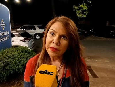 Mujer de cabello largo y suelto, vestida con blusa roja y azul, sostiene un micrófono mientras habla en un ambiente urbano.
