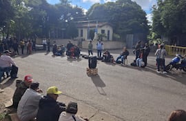 Grupo de personas sentadas en el suelo y sillas, escuchando atentamente en un ambiente exterior con un edificio al fondo.