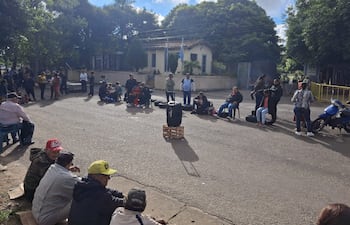 Grupo de personas sentadas en el suelo y sillas, escuchando atentamente en un ambiente exterior con un edificio al fondo.