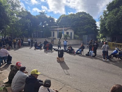 Grupo de personas sentadas en el suelo y sillas, escuchando atentamente en un ambiente exterior con un edificio al fondo.