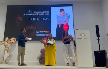 Berta Rojas sosteniendo un galardón en el escenario, vestida de manera elegante, mientras sonríe rodeada de otros presentes.