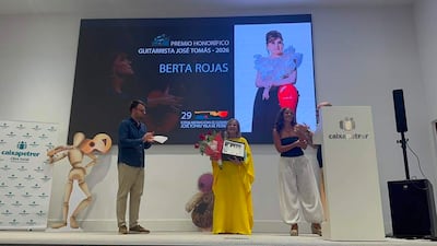 Berta Rojas sosteniendo un galardón en el escenario, vestida de manera elegante, mientras sonríe rodeada de otros presentes.