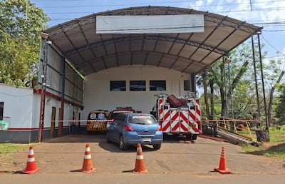 Vehículo azul y camiones de bomberos en un garaje, rodeados de conos naranjas bajo un cobertizo metálico.