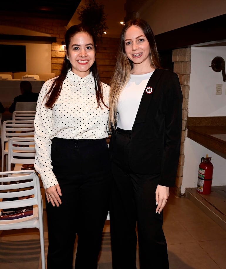 Dos mujeres sonriendo, una en blusa blanca con puntos negros y otra en blusa blanca con saco negro, en un ambiente formal.