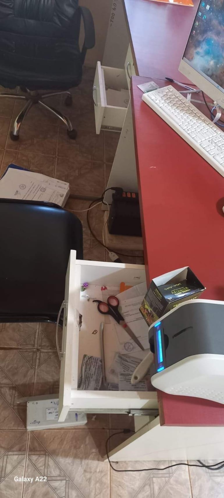 Escritorio rojo con computadora y papeles desordenados en oficina vacía sin personas visibles.