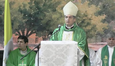 Un obispo con vestimenta clerical verde habla desde un atril, acompañado por dos hombres vestidos en tonos similares en un ambiente solemne.