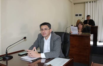 Hombre de cabello corto y canoso en saco gris escucha atentamente. Mujer con cabello largo sostiene documentos. Ambiente formal y sereno.