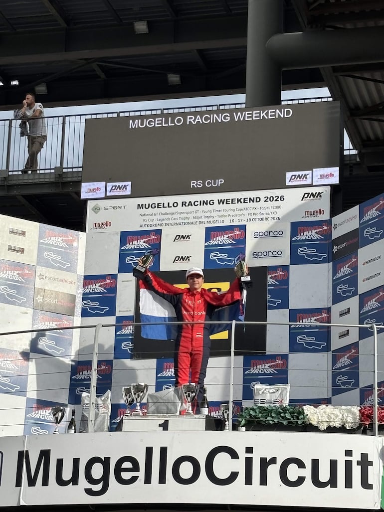 Piloto en traje rojo agita bandera y sostiene trofeo en el podio, con fondo de banderas y trofeos en el circuito de carreras.