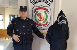 Oficial de policía en uniforme serio junto a persona en capucha negra en un entorno de comisaría.