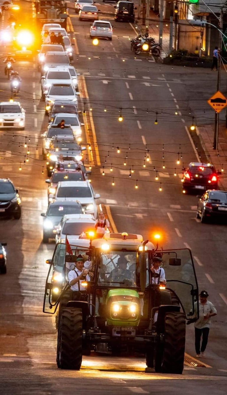 Tractor en desfile con hombre de sombrero y vestimenta formal, rodeado de vehículos y personas al atardecer.