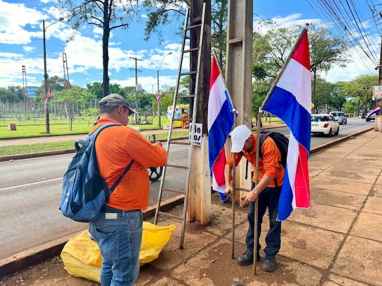 Dos trabajadores de camiseta naranja instalan banderas paraguayas en un entorno verde, enfocados en su tarea mientras un vehículo pasa al fondo.