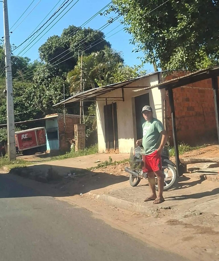 Hombre descalzo con camiseta verde y shorts rojos, apoyado en su bicicleta en una calle con árboles y una vivienda al fondo.