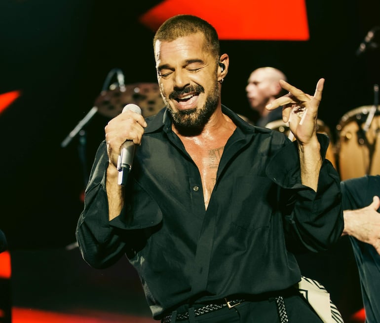Ricky Martin en camisa negra, sosteniendo un micrófono y mostrando felicidad durante su actuación en un evento musical.