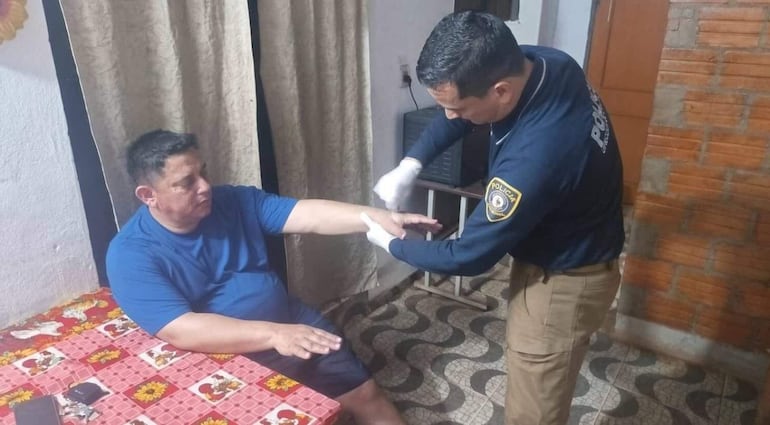 Hombre en uniforme policial venda la muñeca de otro hombre sentado en una mesa con mantel colorido, en un ambiente doméstico.