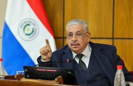 Isaías Fretes en traje oscuro y gafas, gesticulando mientras habla en un ambiente institucional con detalles de la bandera de Paraguay en el fondo.
