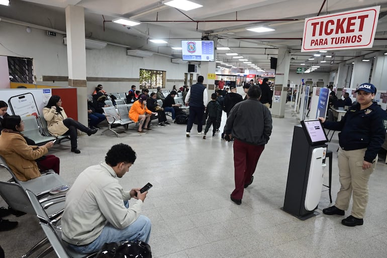 Hombres y mujeres de diversas edades esperan sentados y de pie en una sala moderna, mirando sus teléfonos móviles.
