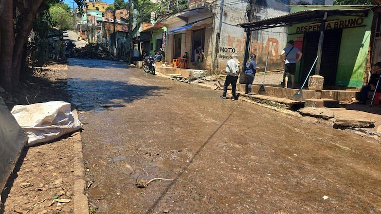 Seis personas trabajan en la calle inundada, usando palas y escobas, con un comercio llamado 'Despenza Bruno' al fondo.