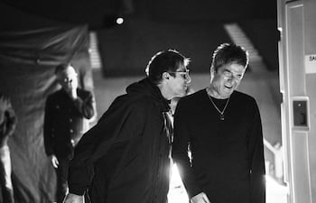 Dos hombres sonriendo y conversando en un ambiente iluminado tenue, con un tercer hombre parcialmente visible al fondo.