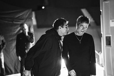Dos hombres sonriendo y conversando en un ambiente iluminado tenue, con un tercer hombre parcialmente visible al fondo.