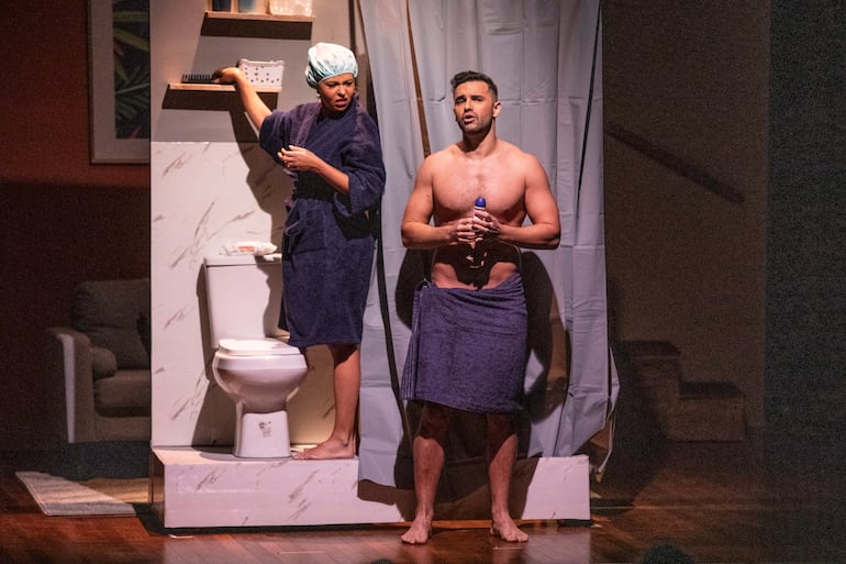 Actriz en toalla azul y gorro de ducha con expresión de sorpresa junto a actor desnudo de la parte superior, en un baño simulado.
