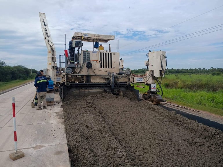 Tres trabajadores en camisetas oscuras y pantalones de trabajo, operan maquinaria pesada en una obra de pavimentación con vegetación al fondo.