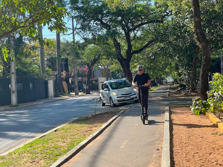 Hombre con casco y ropa oscura monta un patinete eléctrico en vereda arbolada, rodeado de vegetación y con un vehículo estacionado al fondo.