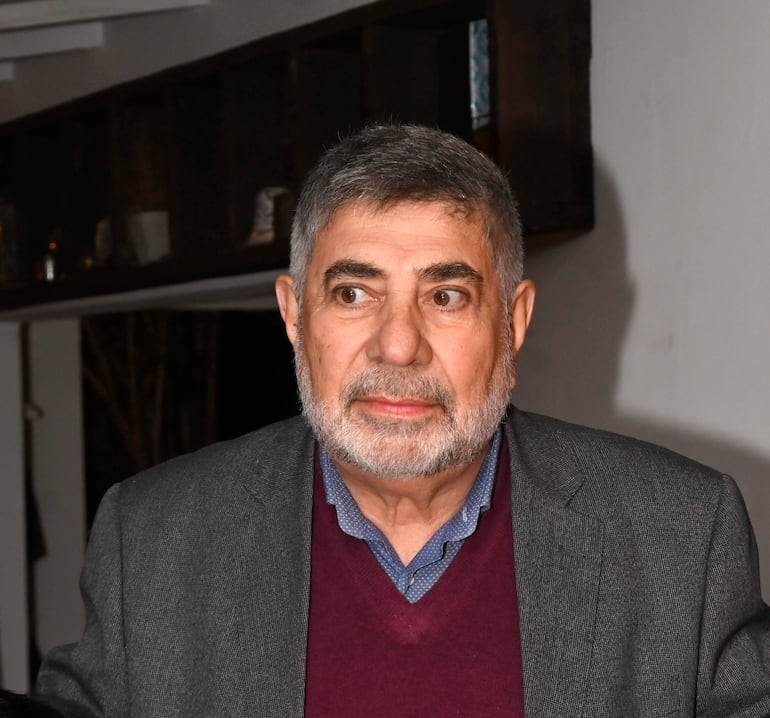 Hombre de mediana edad con barba y cabello canoso, lleva abrigo gris y suéter burdeos, en un ambiente interior.