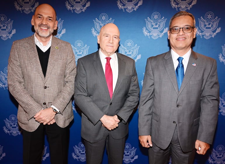 Tres hombres en trajes formales sonríen. Ambiente azul diplomático con símbolos en el fondo.