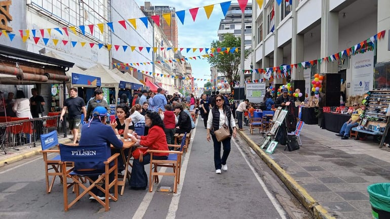 Varias personas conversan y se divierten en una calle adornada, con mesas y comida típica en un ambiente festivo.