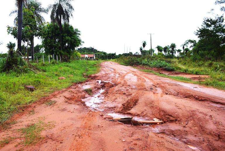 Camino de tierra con surcos y baches, rodeado de vegetación y casas dispersas, bajo un cielo nublado en un entorno rural.