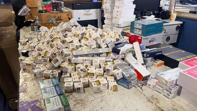 Montón desordenado de cajas de medicamentos en oficina, con impresoras y muebles de fondo.
