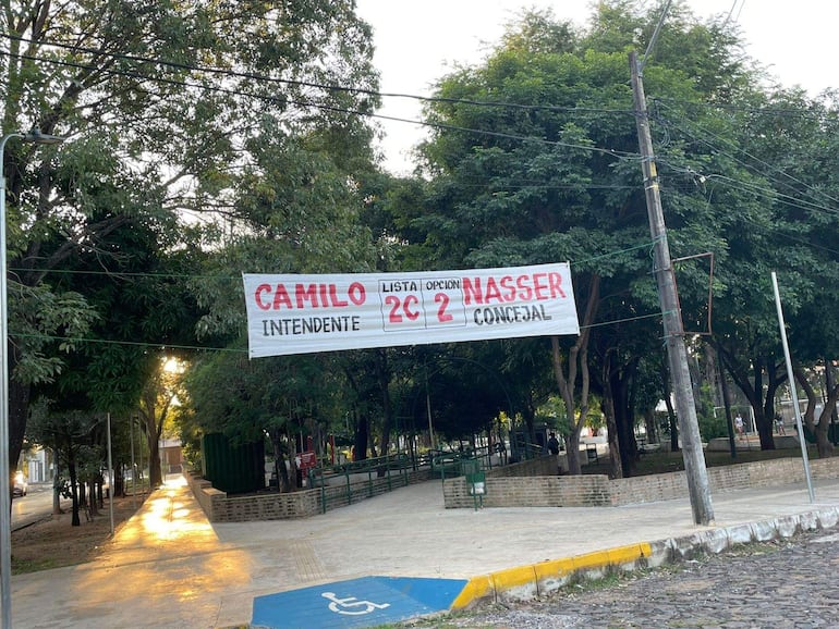 Pasacalle electoral colgado entre postes, con los nombres Camilo Pérez y Nasser Esgaib, en un entorno tranquilo de la plaza.