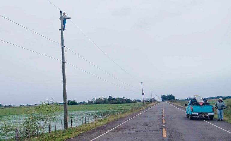 Dos hombres, uno en un poste y otro junto a una camioneta azul, trabajan en un entorno rural con cielo nublado.