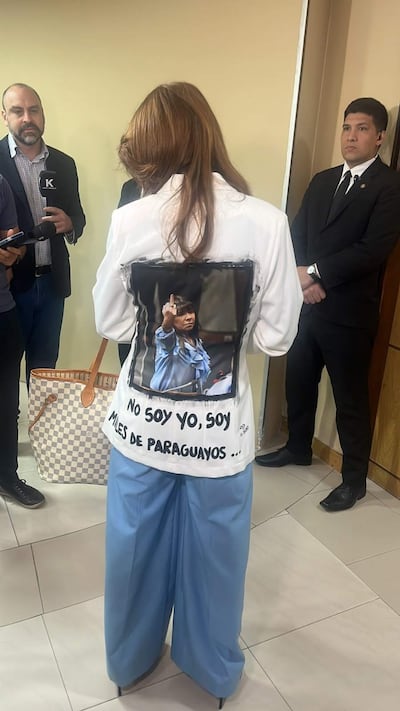 Celeste Amarilla con camisa blanca y pantalones azules, rodeada de personas en un ambiente interior, observa con rostro serio.