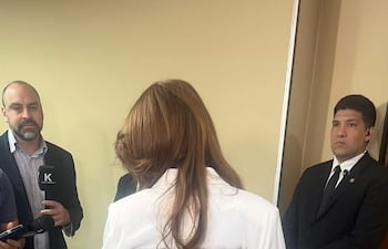 Celeste Amarilla con camisa blanca y pantalones azules, rodeada de personas en un ambiente interior, observa con rostro serio.