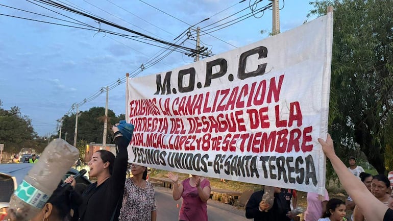 Mujer sostiene un gran cartel en protesta, rodeada de vecinos en ropa casual, en un entorno público con árboles de fondo.