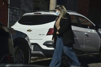 Una mujer con abrigo negro y suéter beige camina por la calle, con mascarilla y cabello largo y rubio, rodeada de vehículos.