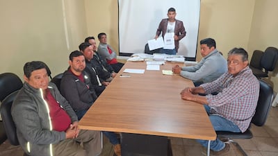 Abel Brítez de pie con documentos, rodeado de hombres sentados en mesa, algunos con expresiones serias, en ambiente laboral.