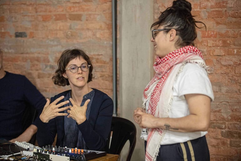 Mujer de blusa azul gesticulando mientras otra mujer, con pañuelo rojo y camiseta blanca, escucha en una sala de reuniones.