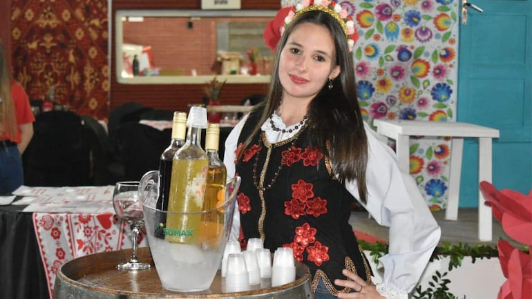 Mujer en vestido negro con flores rojas y diadema de flores, preparando bebidas en una mesa decorada, rodeada de un ambiente festivo.