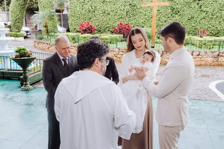 Sacerdote de espaldas en túnica blanca, mujer con bebé en atuendo blanco, hombre mayor y otra persona en un jardín soleado.