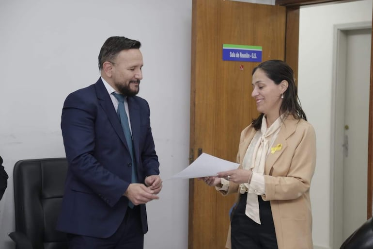 Derlis León en traje oscuro sonríe mientras recibe un documento de la doctora Irene Rocío Benítez Gómez, vestida en blazer claro.