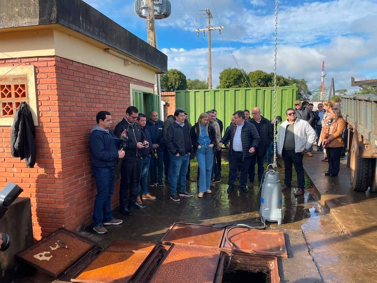 Grupo de personas en acto al aire libre, con un hombre y una mujer rubia en overol al centro, frente a estructura de ladrillo.