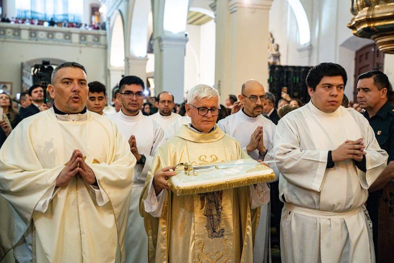 Hombre canoso con gafas y túnica dorada sostiene un objeto, rodeado de hombres con túnicas blancas en un ambiente religioso.