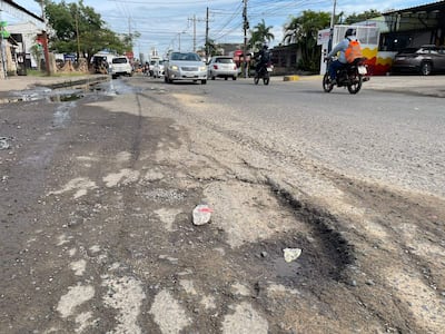 Perspectiva de calle con pavimento agrietado, baches con agua y desechos visibles. Vehículos circulando en la vía.