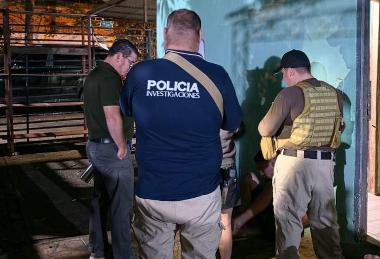 Agentes en uniforme y hombre con camiseta 'POLICÍA INVESTIGACIONES' en operativo, con estructuras metálicas de fondo.