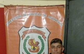 Hombre descalzo en chaqueta oscura y pantalones cortos, serio frente a un cartel policial en la comisaría.