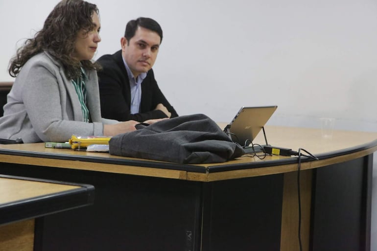 Hernán Rivas, con camisa de cuadros y abrigo negro, escucha atentamente a la mujer de blusa a rayas verdes y abrigo gris en mesa con laptop.