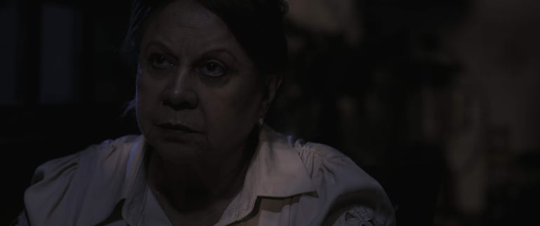 Mujer con cabello oscuro y camisa clara de época, presenta expresión seria en un ambiente sombrío.