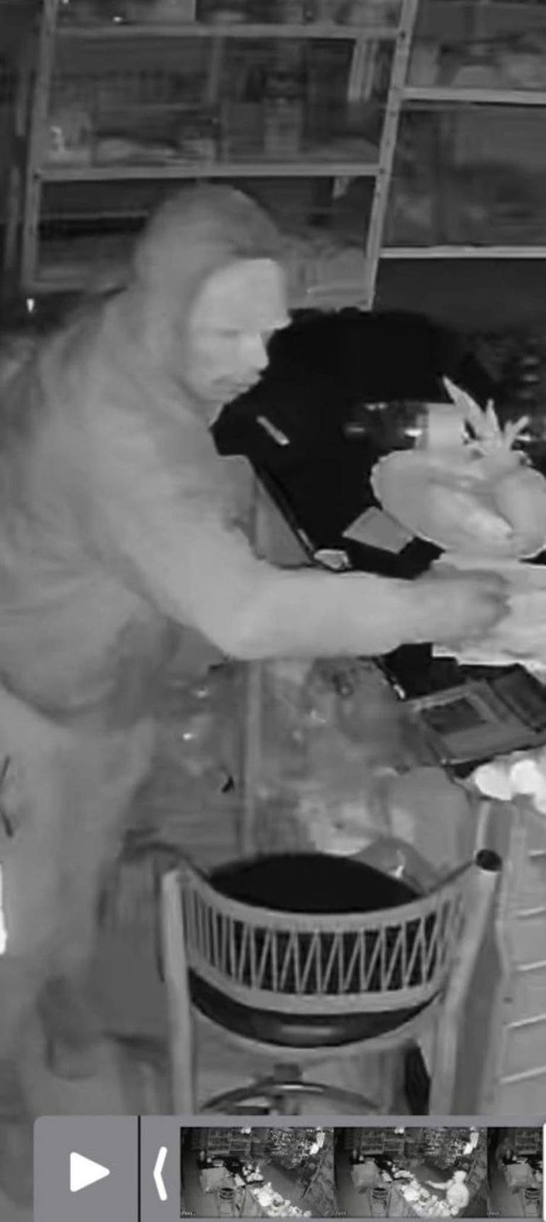 Persona con capucha de pie, inclinada hacia una mesa desordenada en un establecimiento comercial oscuro.